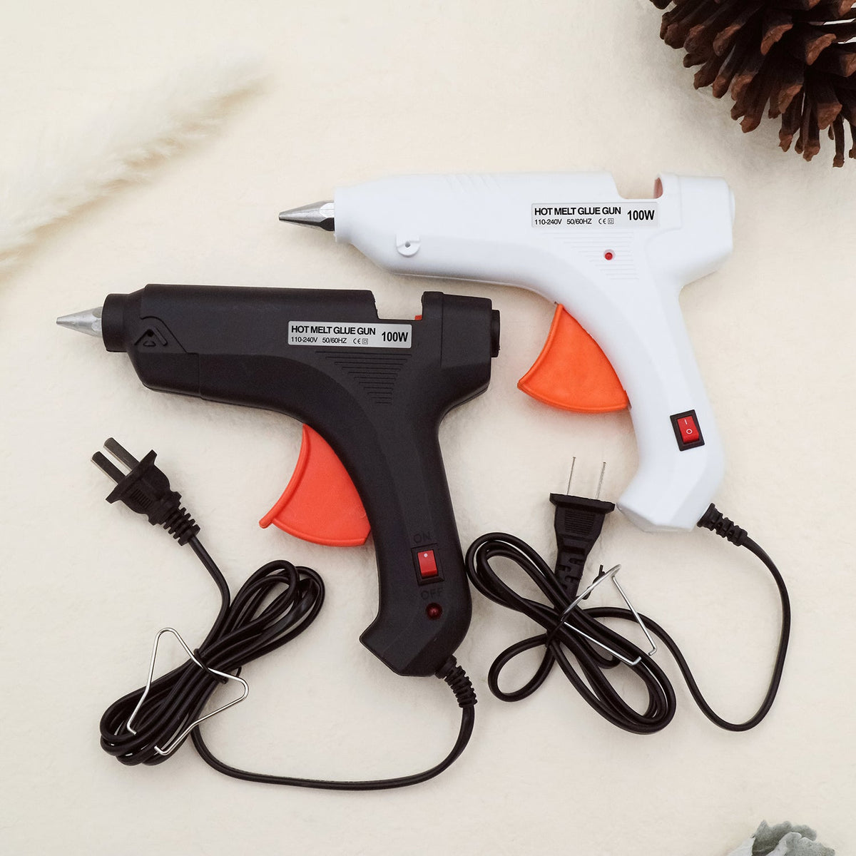 Two electric hot melt glue guns in black and white with orange triggers and attached power cords on a light surface next to a pinecone.